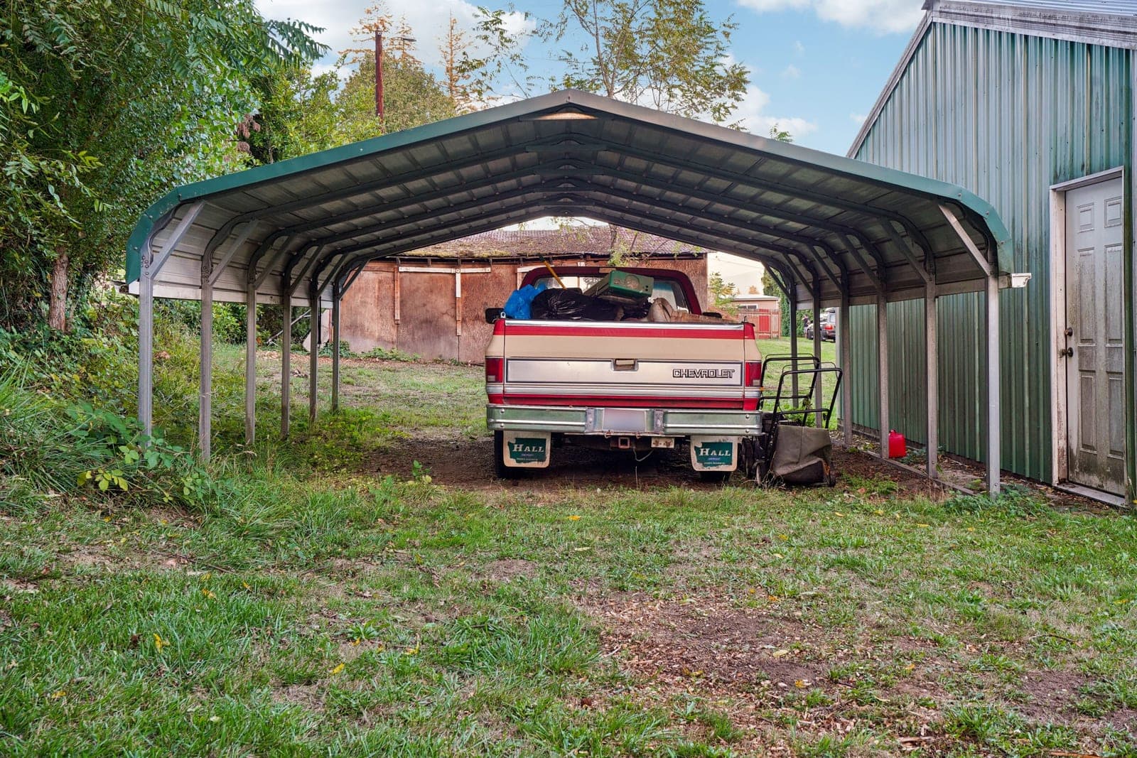 A pickup truck parked under a metal carport in a rural setting