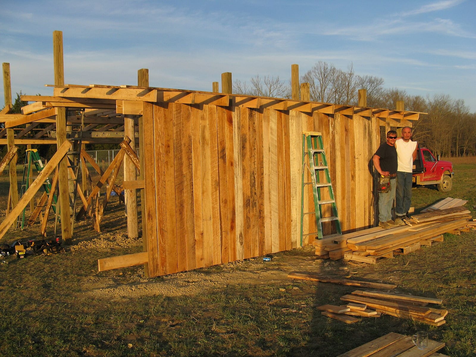 A post-frame building under construction, with bare wooden posts and roof trusses standing against an open sky